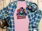 A Garment Graphics pink T-shirt featuring a gorilla in sunglasses and a Gorilla Gangster hat is on a wooden surface, surrounded by a blue and green flannel shirt, headphones, and a camera.