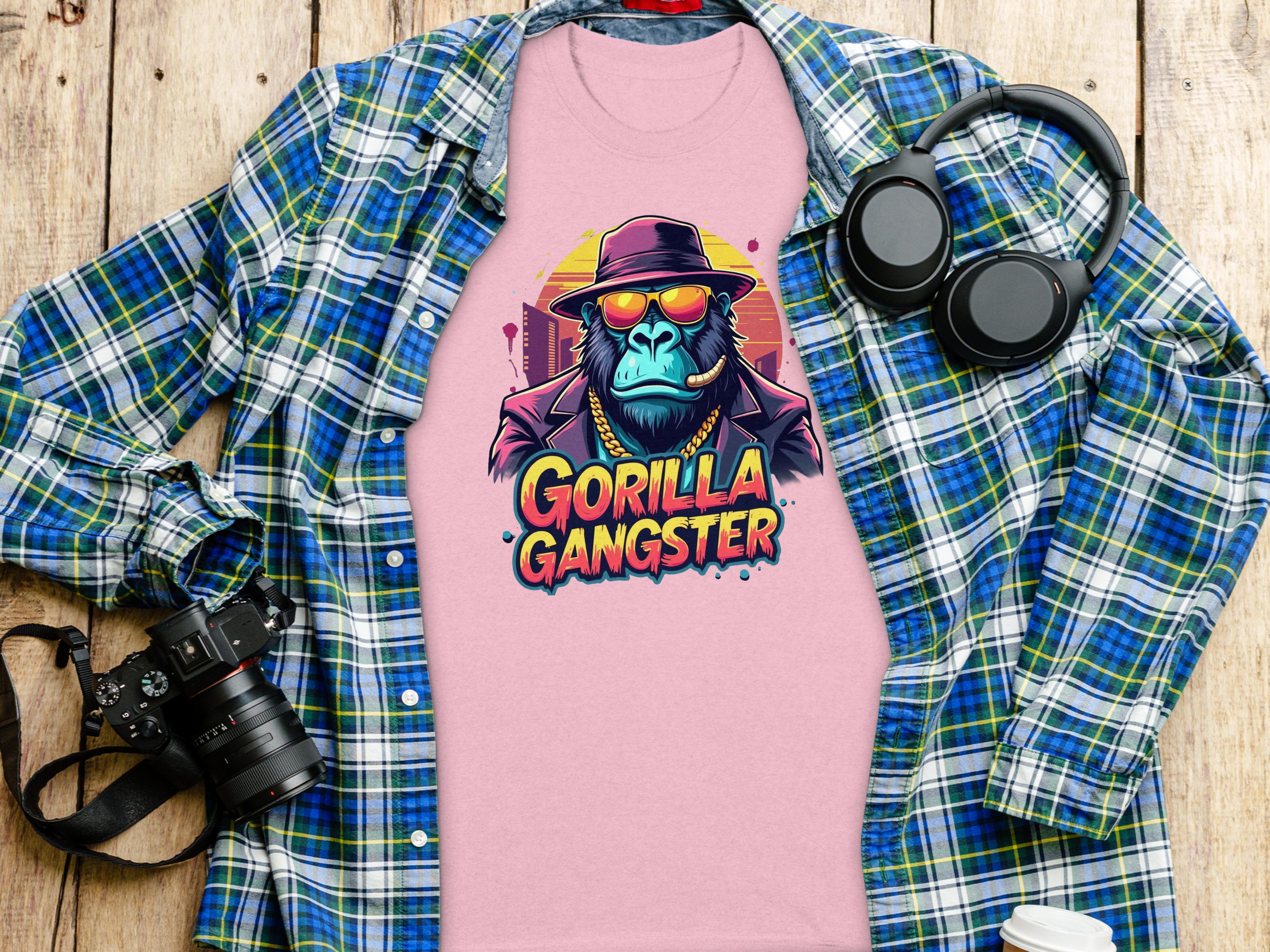 A Garment Graphics pink T-shirt featuring a gorilla in sunglasses and a Gorilla Gangster hat is on a wooden surface, surrounded by a blue and green flannel shirt, headphones, and a camera.