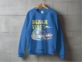 A blue Garment Graphics sweatshirt hangs on a concrete wall, featuring a retro design with surfboards, a wave, and BEACH VIBES, capturing beach fun and classic car nostalgia.