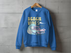 A blue Garment Graphics sweatshirt hangs on a concrete wall, featuring a retro design with surfboards, a wave, and BEACH VIBES, capturing beach fun and classic car nostalgia.