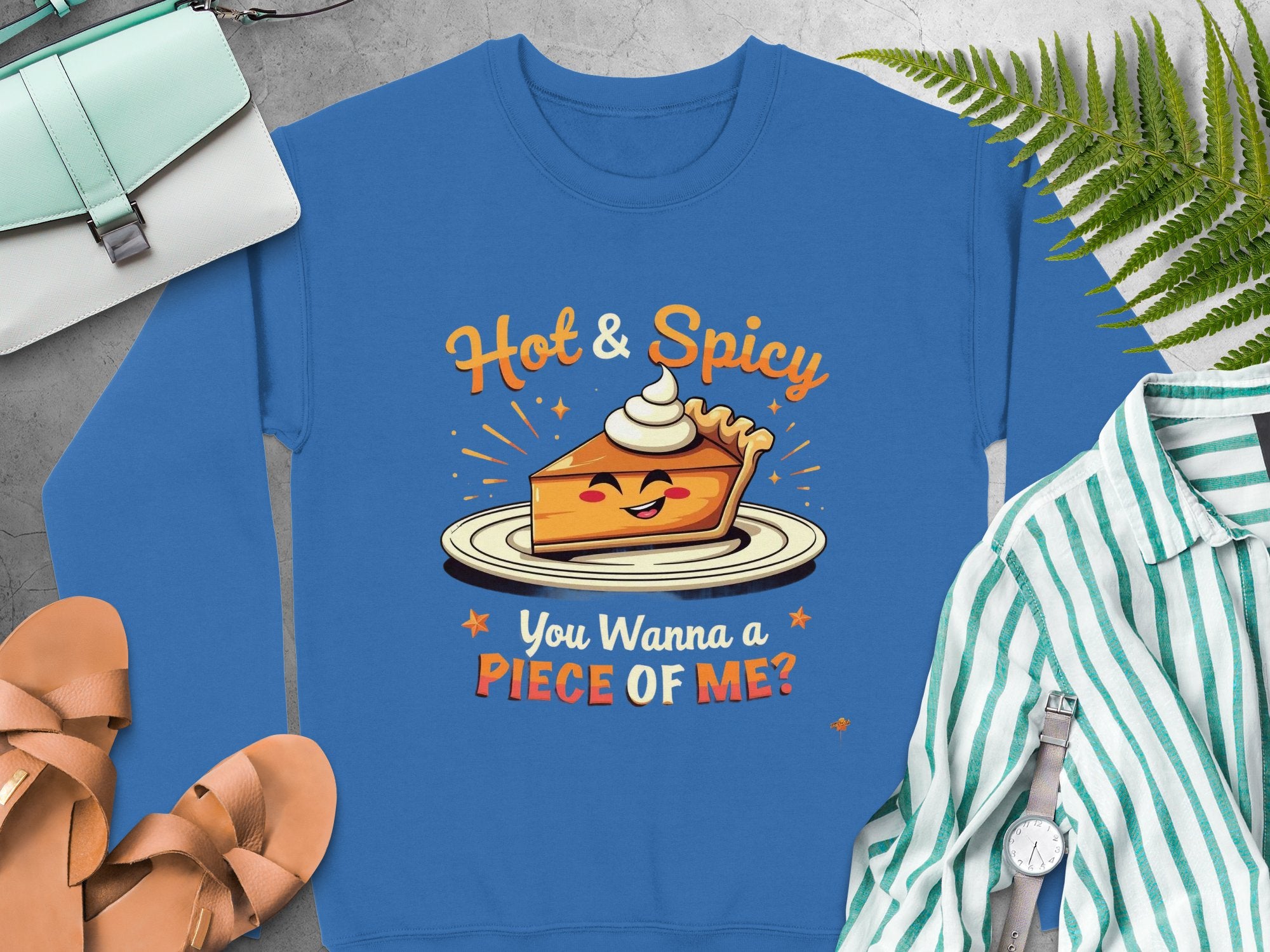 Garment Graphics offers a blue pie graphic sweatshirt with a smiling pie slice and whipped cream, featuring the text Hot & Spicy You Wanna a Piece of Me? This unisex piece is fun and quirky, perfect with sandals, a striped shirt, watch, purse, and green fern leaves.