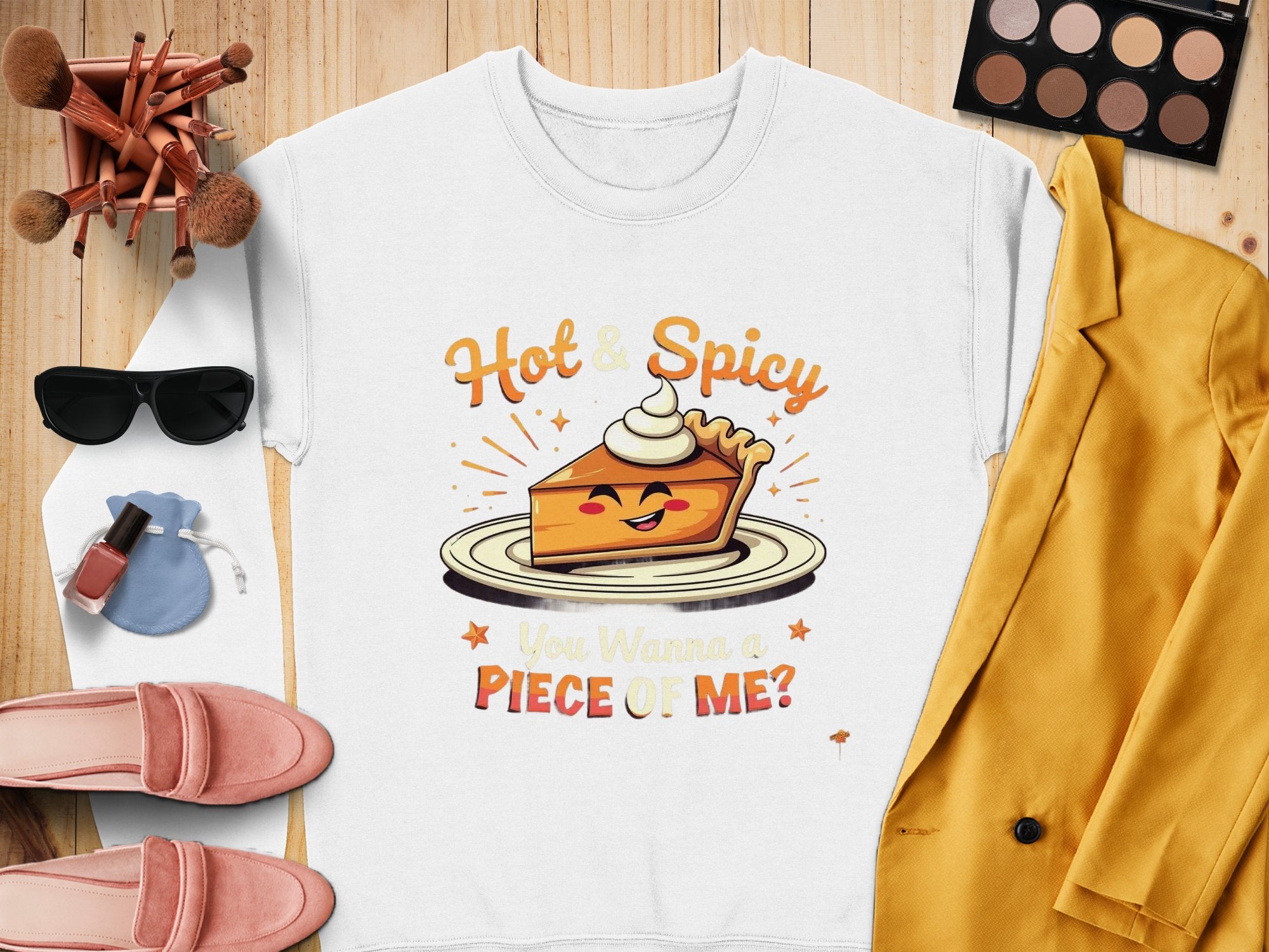 A flat lay showcases a Garment Graphics pie graphic sweatshirt with Hot & Spicy. You Wanna a Piece of Me? text, surrounded by makeup brushes, eyeshadow, sunglasses, blush shoes, pink nail polish, a blue pouch, and a yellow blazer—highlighting this unisex wardrobe staple.