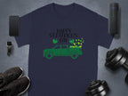 A navy blue Garment Graphics T-shirt features a green pickup truck and shamrocks with Happy St. Patricks Day. Pictured with a smartphone, smartwatch, wireless speaker, shoes, and dumbbells on a gray background for stylish Irish pride.