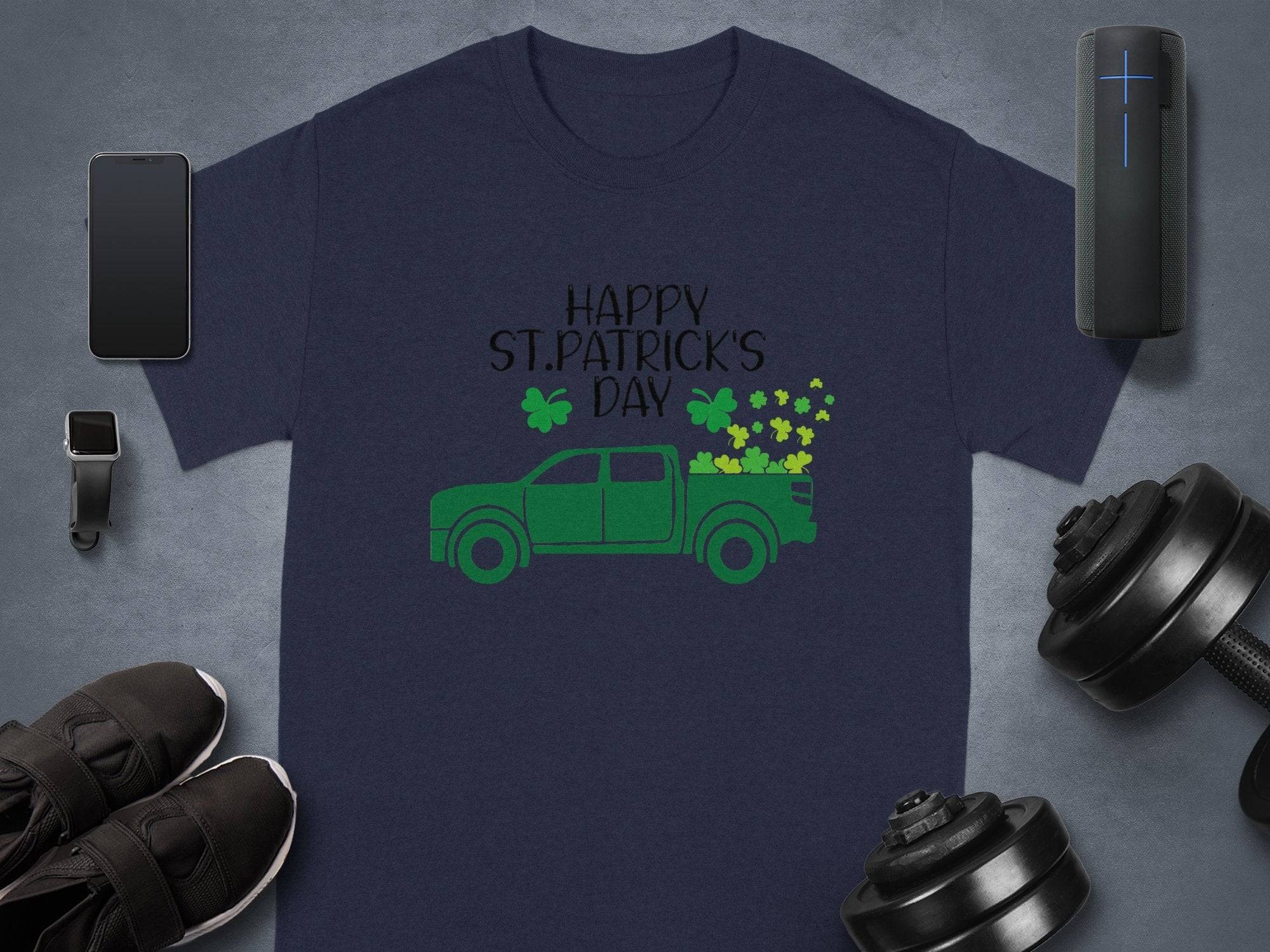 A navy blue Garment Graphics T-shirt features a green pickup truck and shamrocks with Happy St. Patricks Day. Pictured with a smartphone, smartwatch, wireless speaker, shoes, and dumbbells on a gray background for stylish Irish pride.