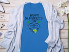 The Garment Graphics St. Patricks Day graphic tee features Happy St. Patricks Day text, green and yellow shamrocks, and a heart on a blue shirt. It’s styled with a light gray cardigan on a wooden surface accented by white flowers and accessories.