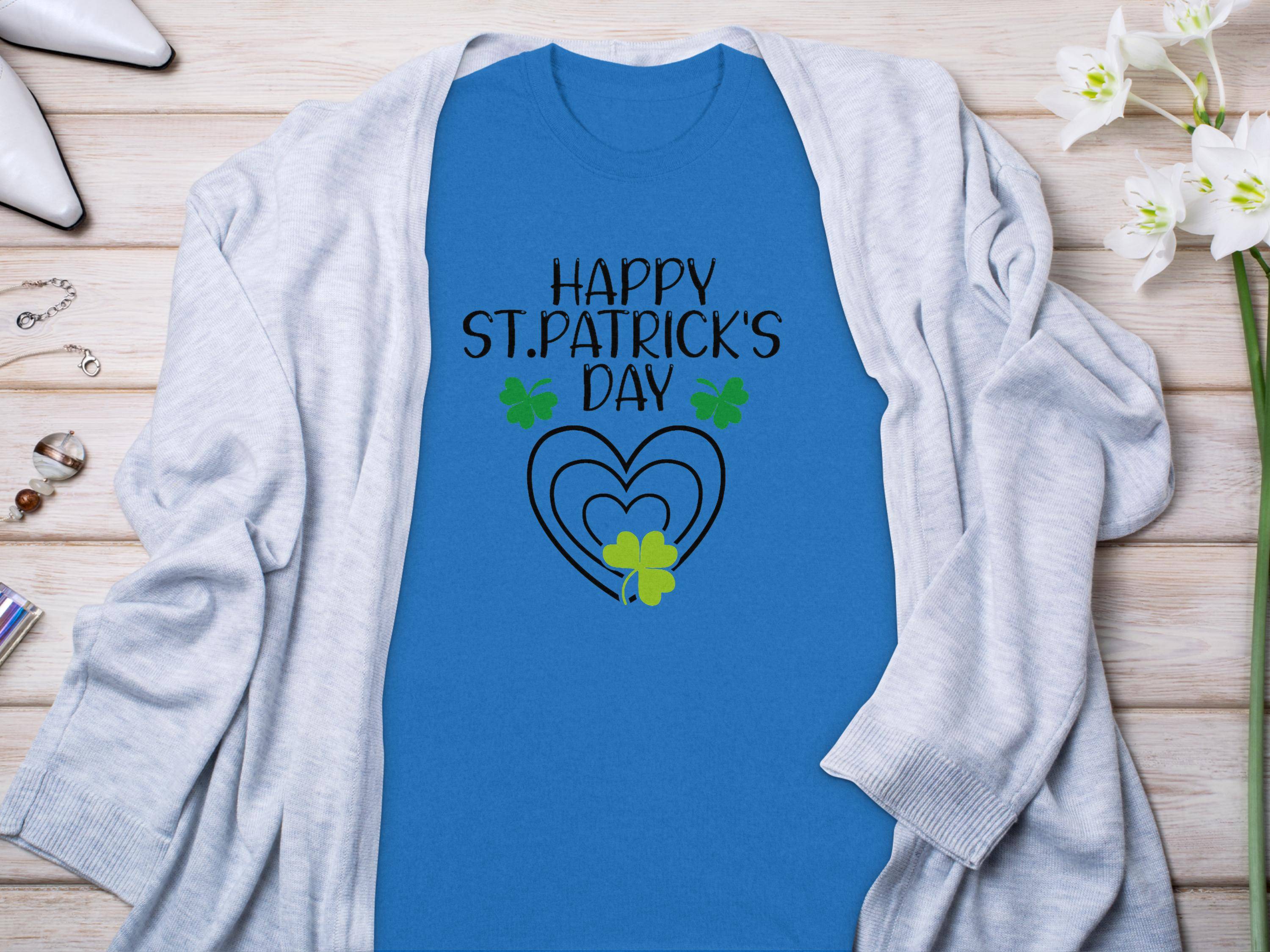 The Garment Graphics St. Patricks Day graphic tee features Happy St. Patricks Day text, green and yellow shamrocks, and a heart on a blue shirt. It’s styled with a light gray cardigan on a wooden surface accented by white flowers and accessories.