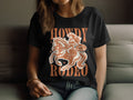 A person sits on a couch wearing a black Garment Graphics T-shirt, featuring a rodeo scene illustration and Howdy Rodeo in bold. This womens vintage-style tee with its brown theme is ideal for rodeo fans.