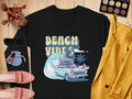 A black shirt from Garment Graphics exudes retro vibes with its Beach Vibes design featuring a classic car, surfboards, waves, and palm trees. Its styled with makeup brushes, an eyeshadow palette, sunglasses, a yellow jacket, pink shoes, and lipsticks on a wooden surface.