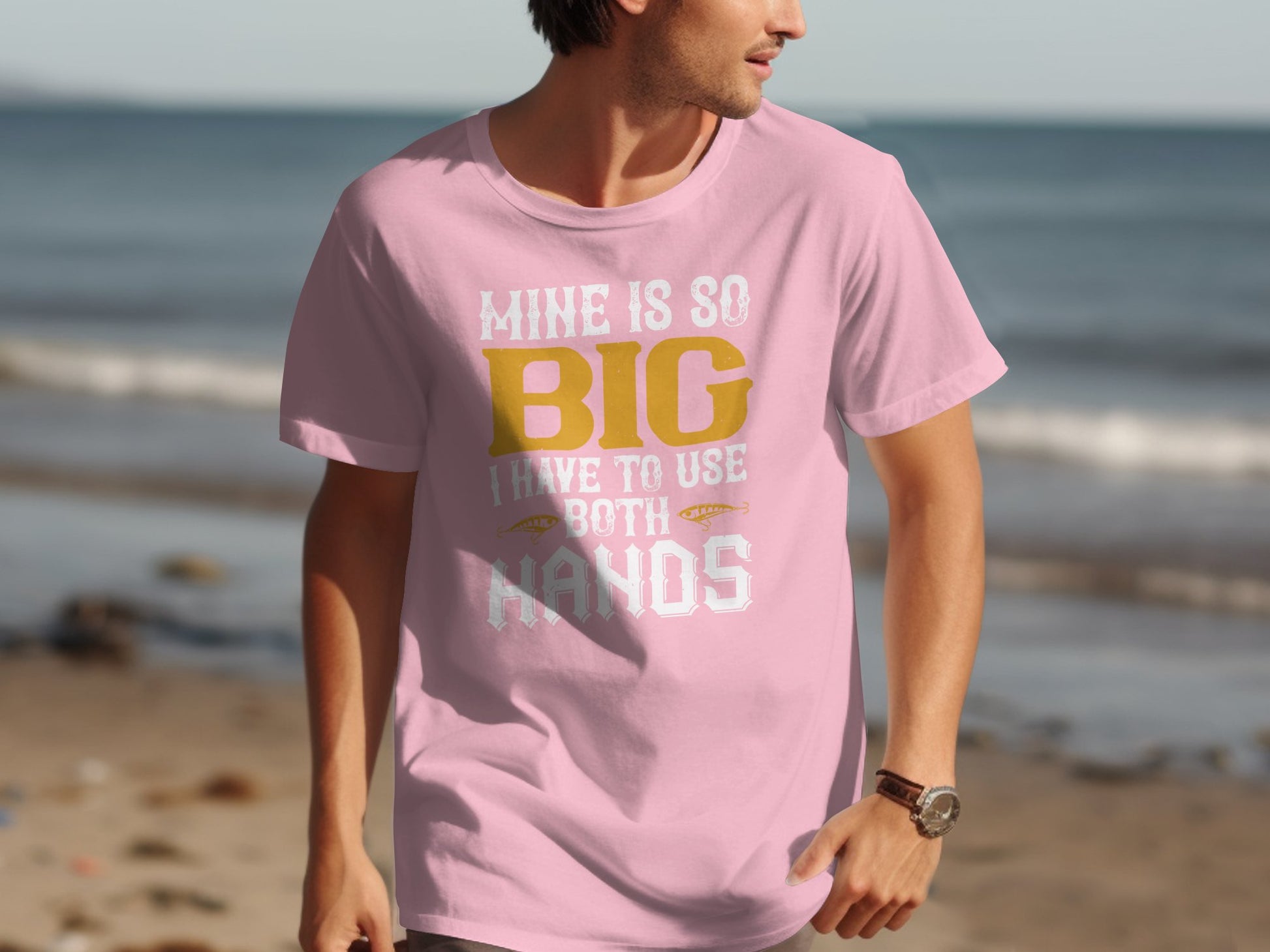 On a beach, a man wears a pink Garment Graphics fishing-themed tee, featuring yellow and white text: Mine is so big I have to use both hands, adding humor for fishing fans. The ocean and sand backdrop enhance its playful design.