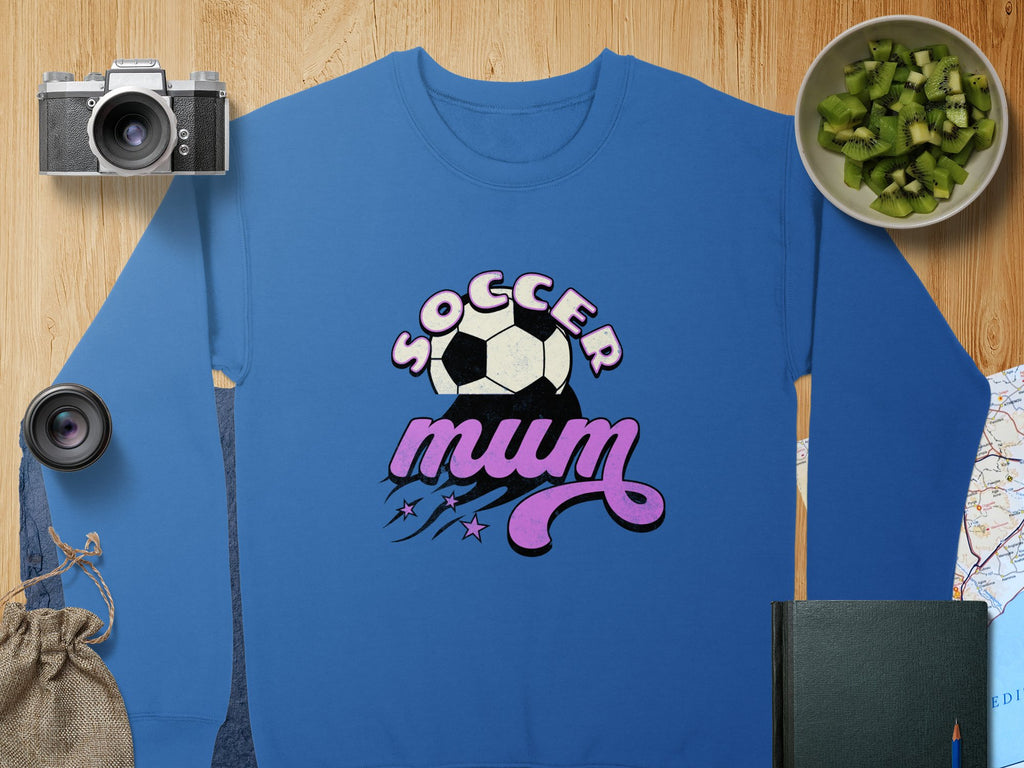 A blue Garment Graphics sweatshirt reads Soccer Mum with a soccer ball and pink stars in a vintage style. Nearby are a camera, lens, small bag, bowl of kiwi slices, book, and part of a map on a wooden surface.