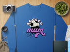 A blue Garment Graphics sweatshirt reads Soccer Mum with a soccer ball and pink stars in a vintage style. Nearby are a camera, lens, small bag, bowl of kiwi slices, book, and part of a map on a wooden surface.