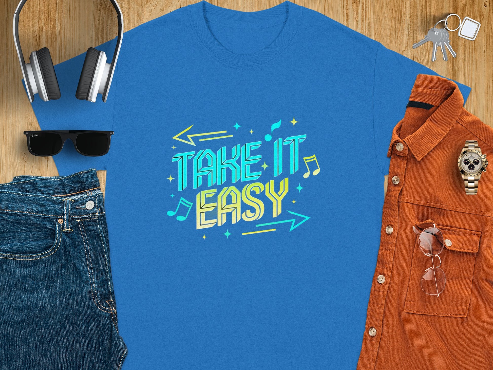 A vibrant Take It Easy T-shirt from Garment Graphics, featuring musical notes and arrows, lies on a wooden surface. Surrounding it are folded jeans, trendy sunglasses, headphones, keys, a sleek watch, and an eye-catching orange button-up shirt.