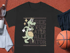 Garment Graphics black T-shirt is ideal for casual wear, showcasing a cartoon alligator spinning a basketball with See You Later Alligator printed behind it. It is stylishly paired with a water bottle, towel, and basketball.