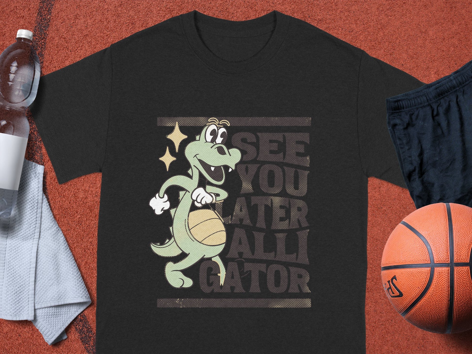 Garment Graphics black T-shirt is ideal for casual wear, showcasing a cartoon alligator spinning a basketball with See You Later Alligator printed behind it. It is stylishly paired with a water bottle, towel, and basketball.