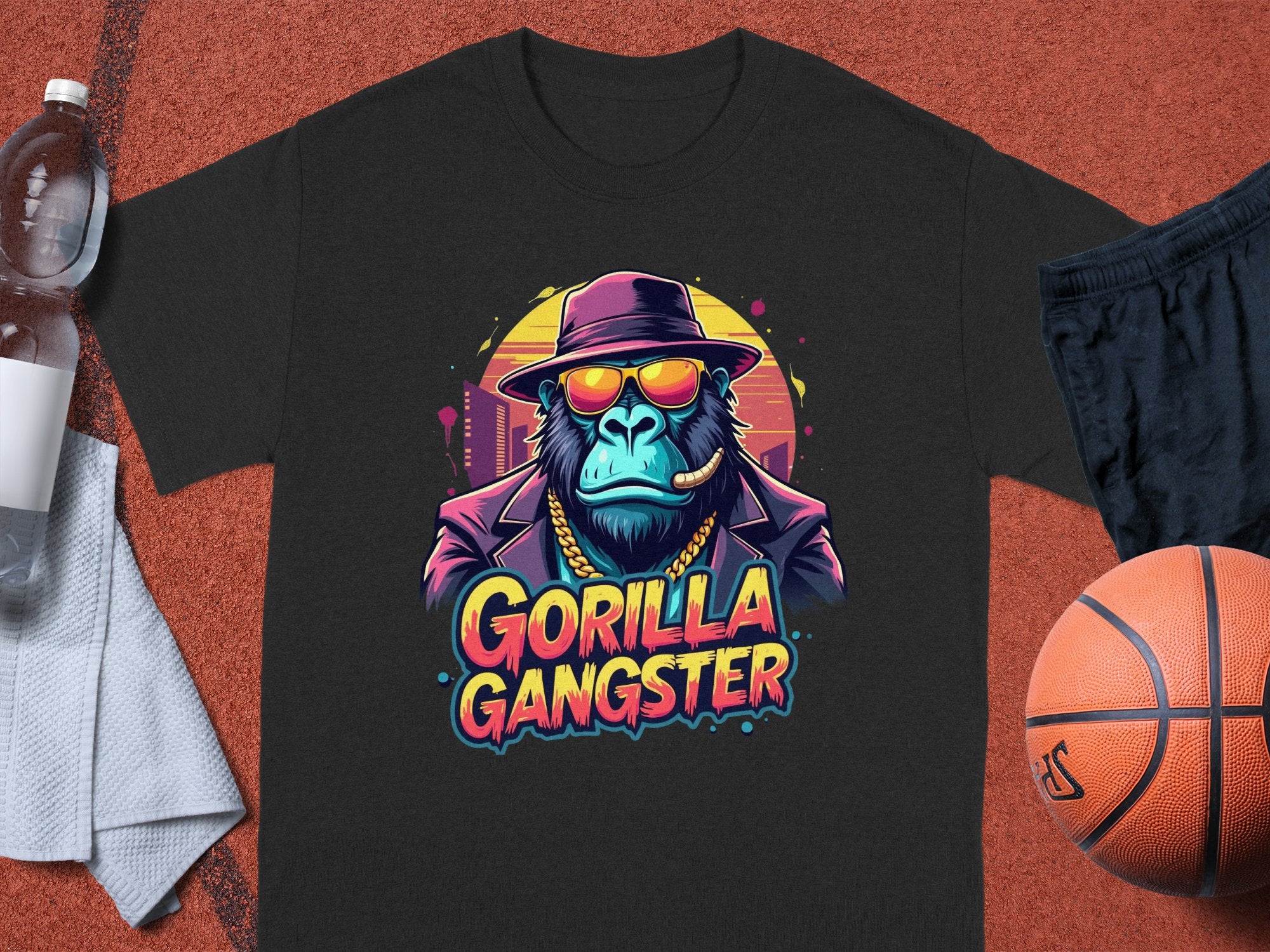 Garment Graphics black T-shirt features a gorilla in a hat, sunglasses, gold chain, cigar, with GORILLA GANGSTER text. Its displayed on a sports court beside a basketball, water bottle, and white towel for a bold look.