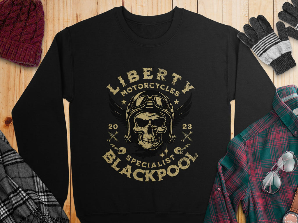 On a wooden surface, a black Garment Graphics sweatshirt features a skull with goggles and helmet, epitomizing the biker vibe. The text reads Liberty Motorcycles Specialist Blackpool 2023. Nearby are accessories: beanie, gloves, plaid shirt, and sunglasses.