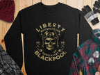 On a wooden surface, a black Garment Graphics sweatshirt features a skull with goggles and helmet, epitomizing the biker vibe. The text reads Liberty Motorcycles Specialist Blackpool 2023. Nearby are accessories: beanie, gloves, plaid shirt, and sunglasses.