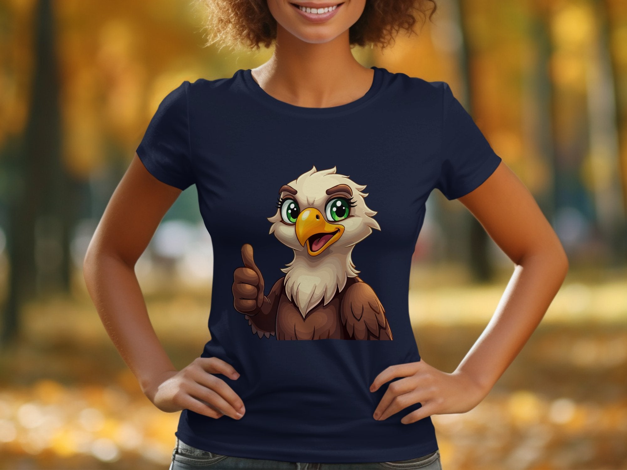 Outdoors, a person wearing Garment Graphics navy cartoon T-shirt featuring an eagle giving a thumbs up stands against a backdrop of blurred autumn trees. Smiling and with hands on hips, the scene is perfect for animal lovers.