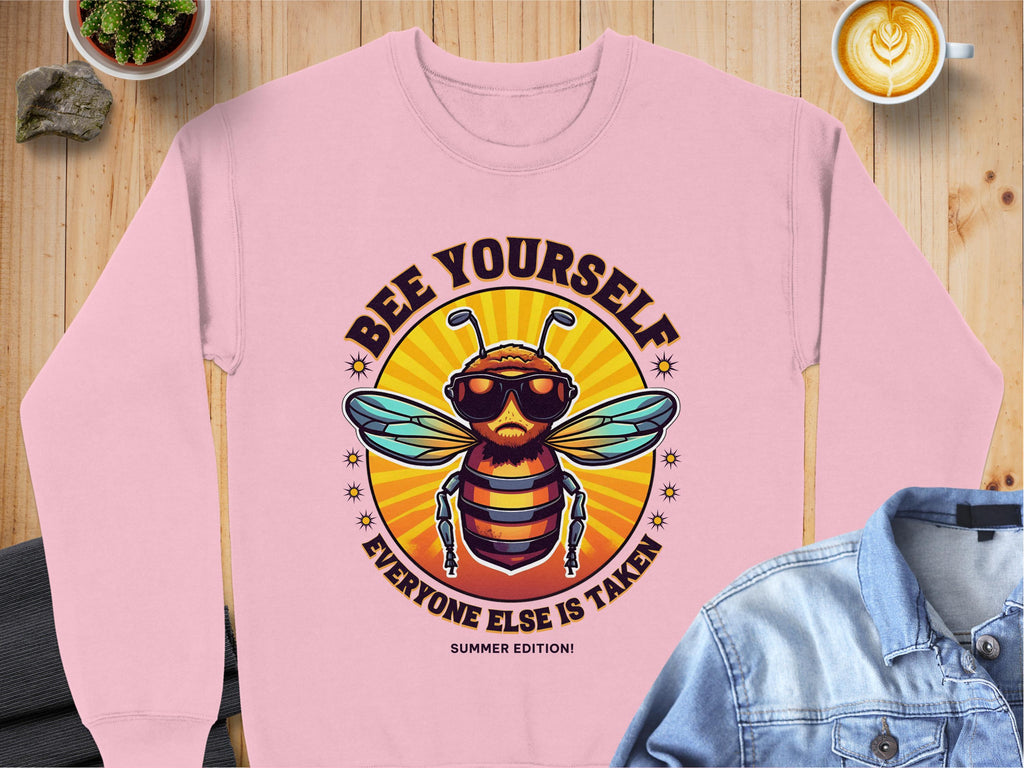 This chic Garment Graphics pink sweatshirt features a cartoon bee in sunglasses with the slogan Bee Yourself, Everyone Else Is Taken! Ideal for summer, it offers unisex sizing and is styled with a denim jacket and latte on a wooden surface.