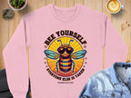 This chic Garment Graphics pink sweatshirt features a cartoon bee in sunglasses with the slogan Bee Yourself, Everyone Else Is Taken! Ideal for summer, it offers unisex sizing and is styled with a denim jacket and latte on a wooden surface.