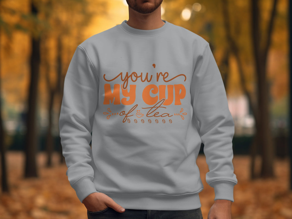 A person wears a cozy light gray Garment Graphics sweatshirt, showcasing Youre My Cup of Tea in vibrant orange. Set against blurred golden autumn leaves, its ideal for tea enthusiasts who cherish seasonal warmth amidst a forest backdrop.