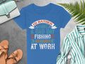 A blue fishing T-shirt by Garment Graphics, ideal for enthusiasts, lies on concrete and reads, Id rather have a bad day fishing than a good day at work, in bold colors. Nearby are brown sandals, a striped shirt, watch, foliage, and green handbag—all perfect for womens sizing.