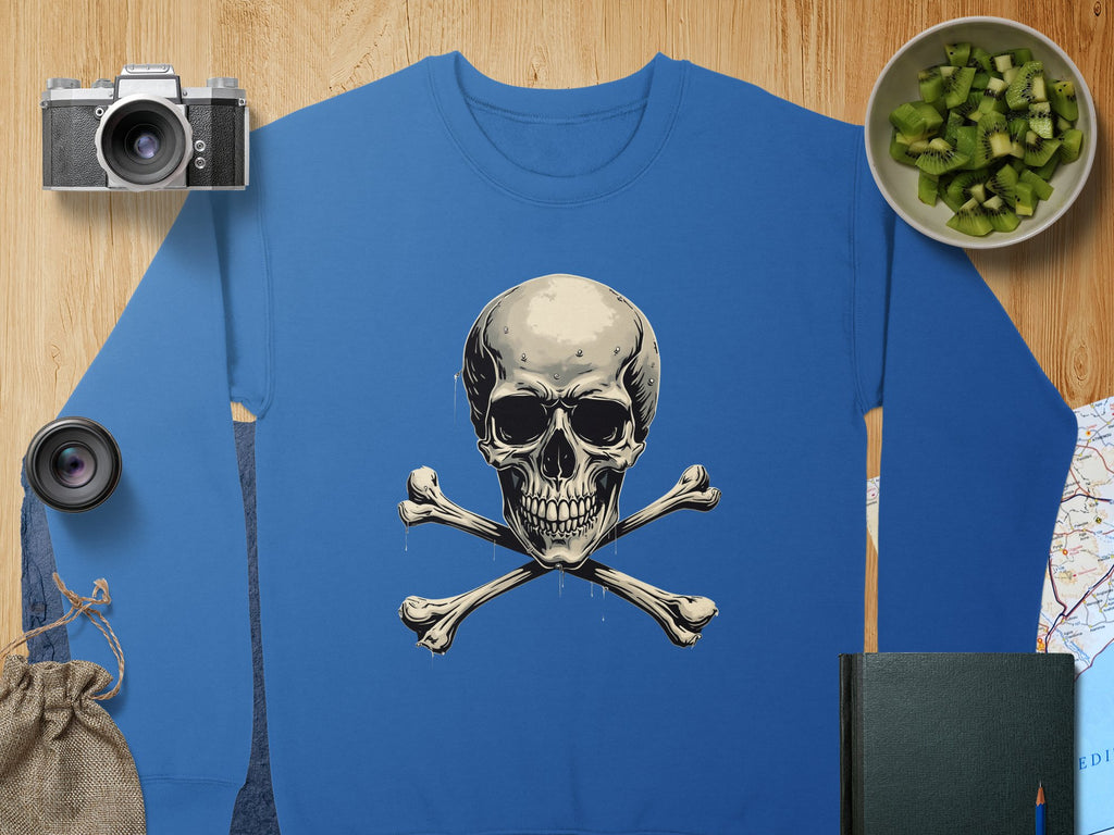 A Garment Graphics blue sweatshirt featuring a skull and crossbones design rests on wood. Its surrounded by a camera, lens, green fruit in a bowl, burlap sack, closed book, and part of a map. Available in unisex sizes for the daring.