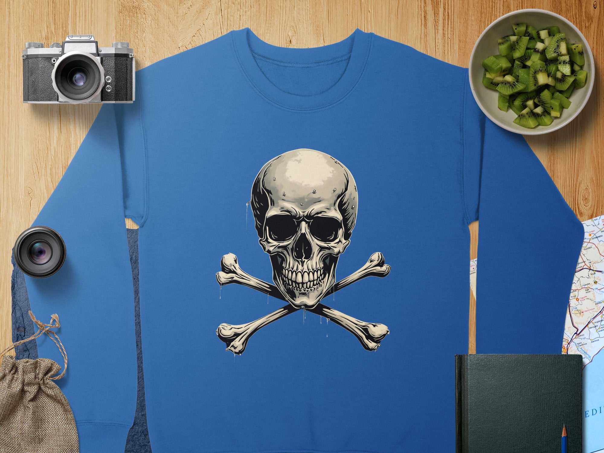 A Garment Graphics blue sweatshirt featuring a skull and crossbones design rests on wood. Its surrounded by a camera, lens, green fruit in a bowl, burlap sack, closed book, and part of a map. Available in unisex sizes for the daring.