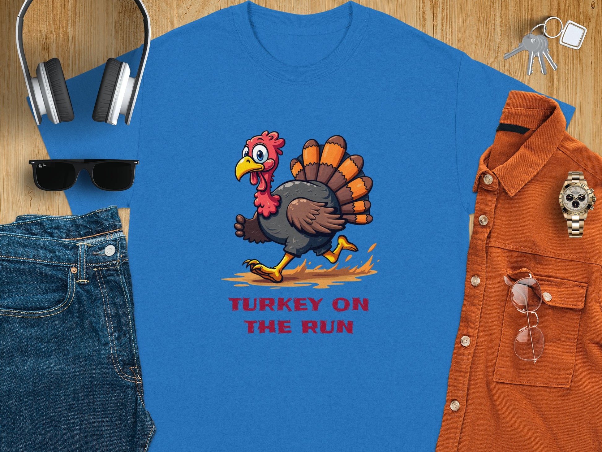 The Garment Graphics classic fit blue t-shirt showcases a humorous turkey graphic with the text Turkey On The Run. Its paired with headphones, a baseball cap, keys, sunglasses, denim jeans, an orange-brown button-up shirt, and a watch.