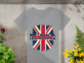This striking gray T-shirt by Garment Graphics is made of 100% cotton and displays the Union Jack with the text Dont Blame Me, I Didnt Vote For Him. Its set on concrete, accented by vibrant red and yellow flowers, and lush green leaves.