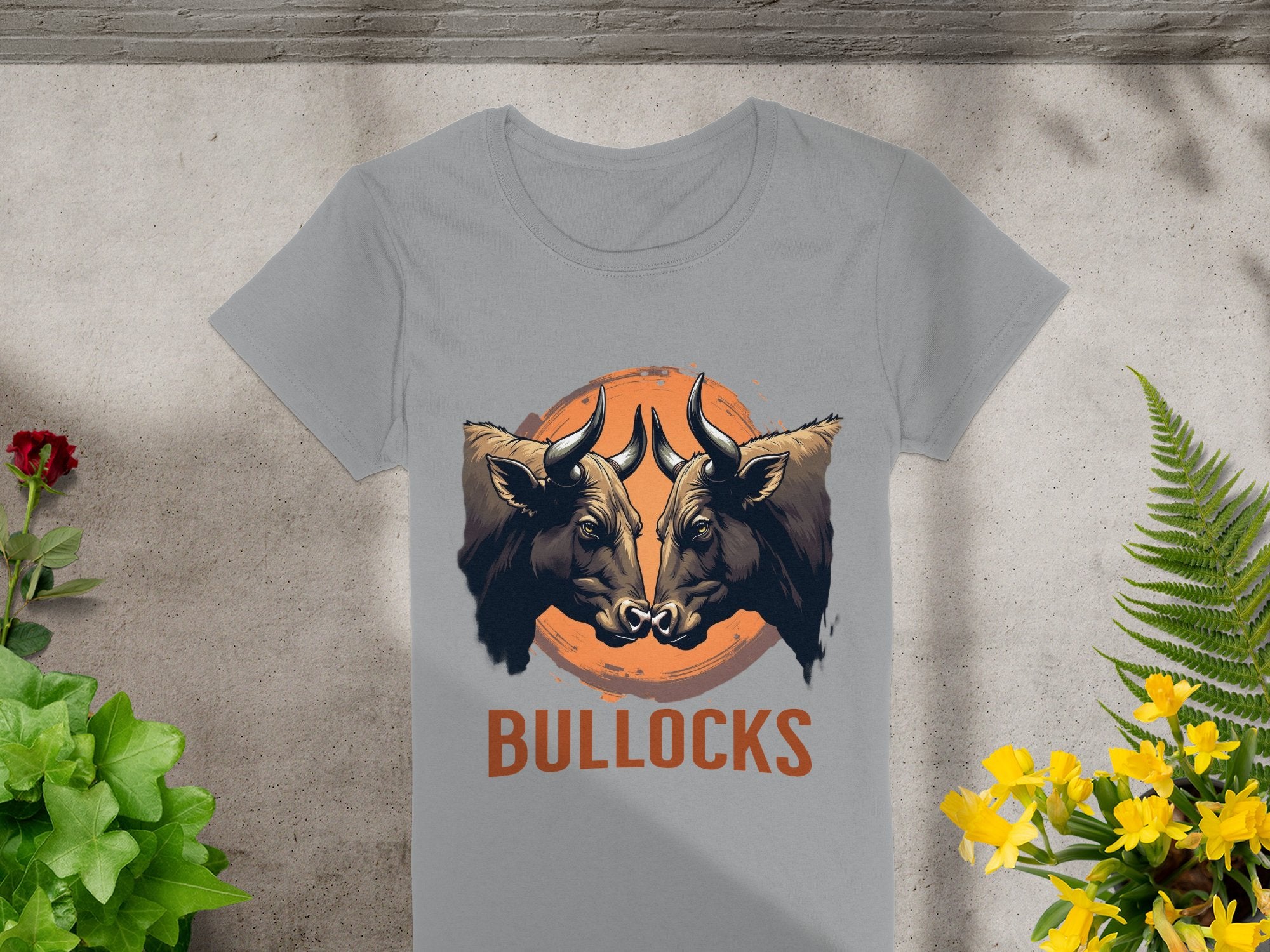 Garment Graphics offers a gray t-shirt featuring striking graphics of two bulls facing each other over an orange circle, with BULLOCKS below. Ideal for casual wear, this shirt is displayed on concrete surrounded by green plants and yellow flowers.