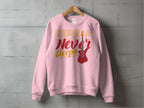 A vibrant pink Garment Graphics sweatshirt hangs on a wooden hanger against a concrete wall, ideal for music fans. It features Music Never Sleeps in bold and showcases a striking red electric guitar graphic on the right side.