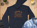 A Garment Graphics black hoodie with This Day is Powered by Coffee in decorative font rests on wood, surrounded by jeans, a heart-detailed latte, a suit jacket, and a small potted plant—a delightful scene for coffee lovers.