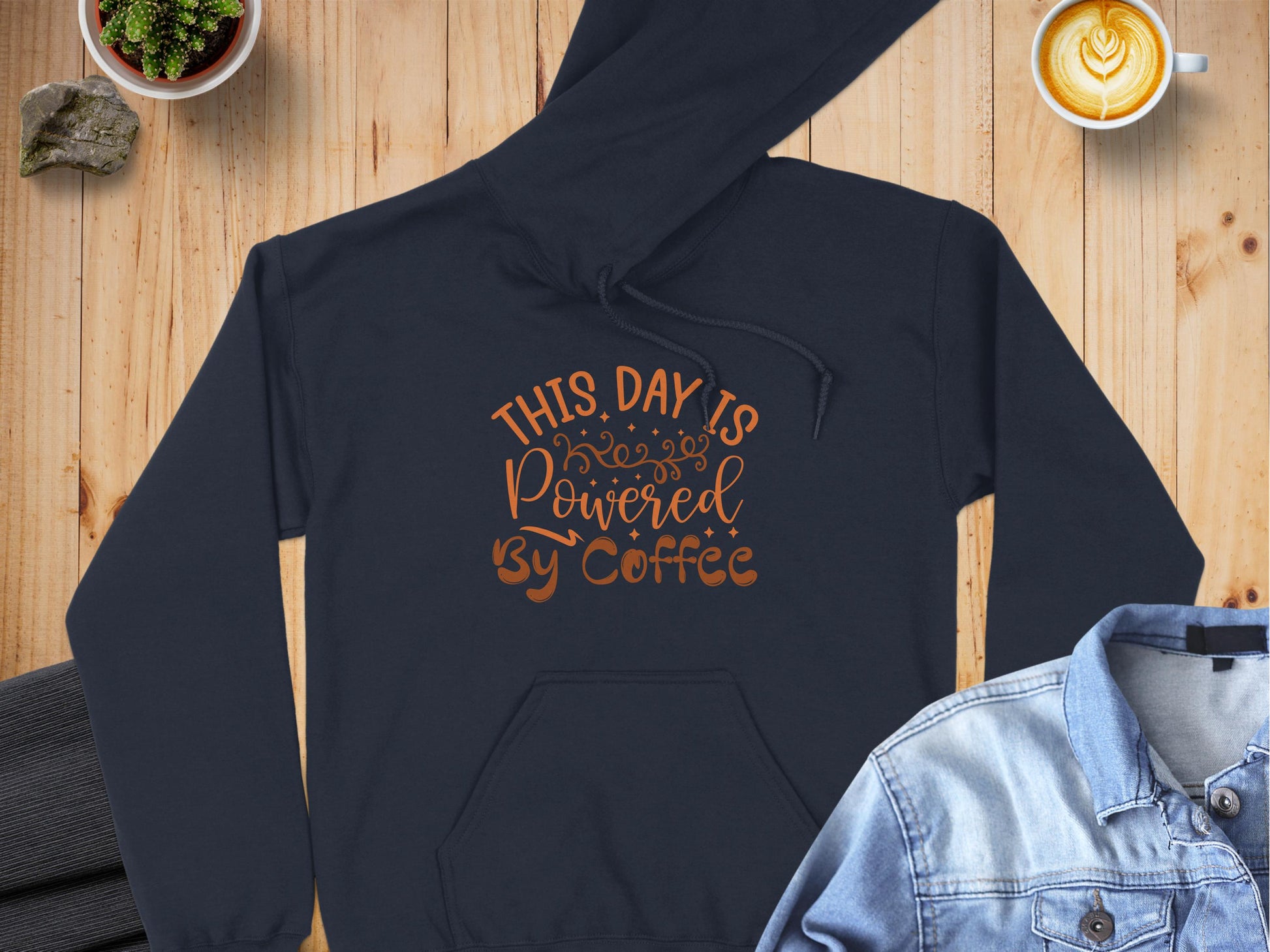 A Garment Graphics black hoodie with This Day is Powered by Coffee in decorative font rests on wood, surrounded by jeans, a heart-detailed latte, a suit jacket, and a small potted plant—a delightful scene for coffee lovers.