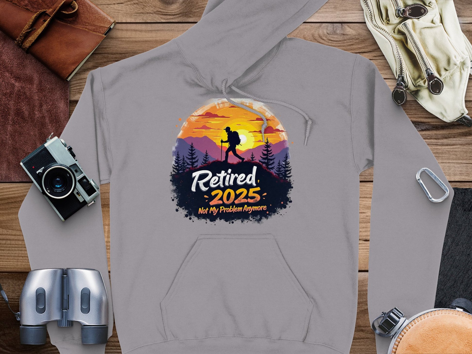 A gray hiking hoodie by Garment Graphics features a silhouette with a pole against a sunset mountain. It reads, Retired 2025: Not My Problem Anymore and is laid on wood, surrounded by travel essentials reflecting adventure and freedom.