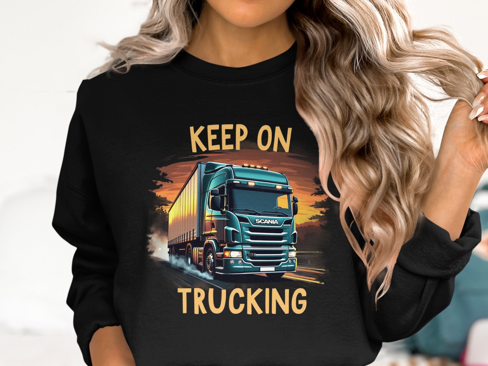 A person with wavy hair sports a Graphic Sweatshirt from Garment Graphics, showcasing a teal semi-truck. The black sweatshirt, ideal for truck enthusiasts, is emblazoned with KEEP ON TRUCKING above and below the image.