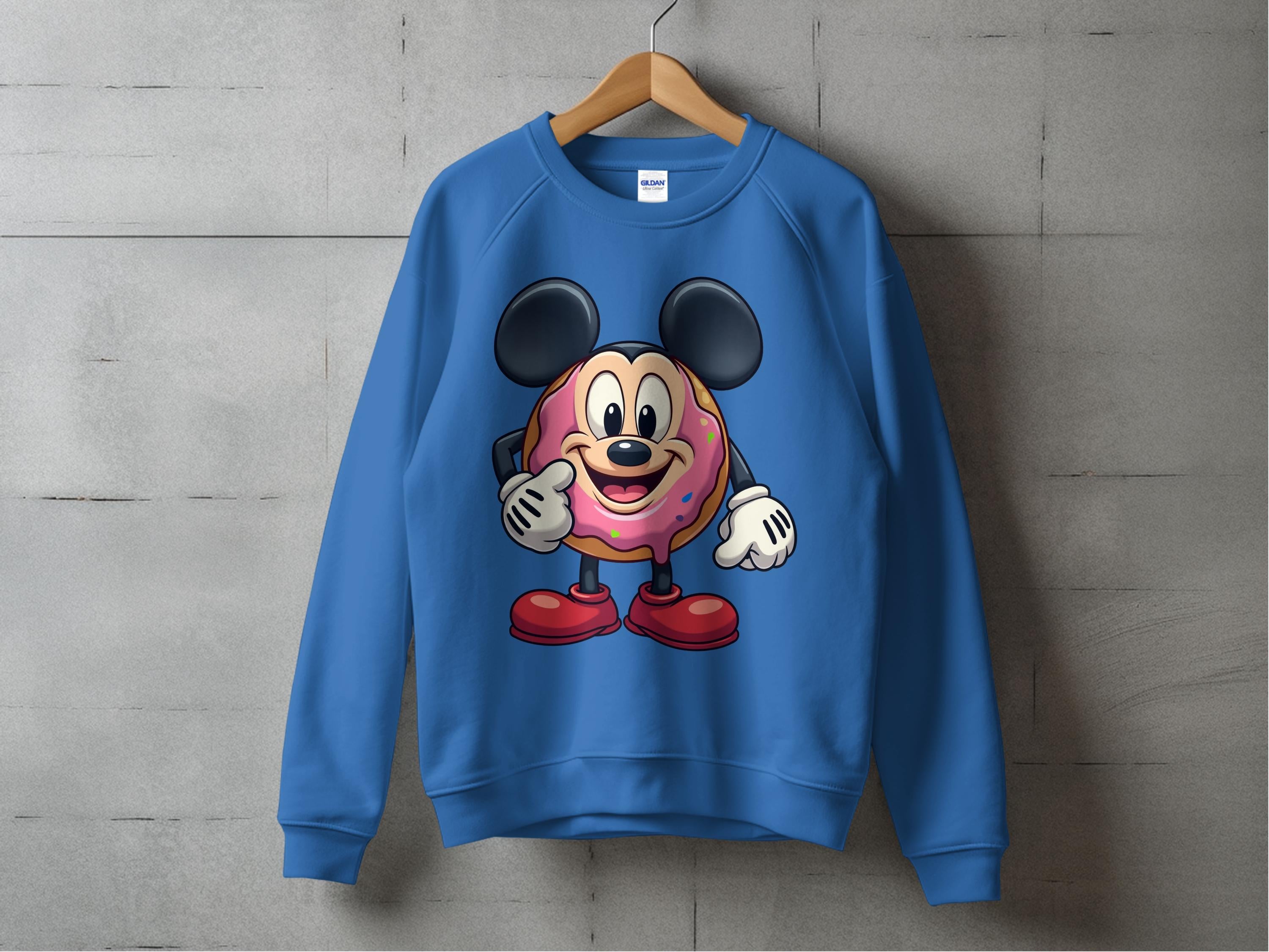 A unisex blue sweatshirt from Garment Graphics hangs on a wooden hanger against a gray concrete wall, featuring a cartoon donut character with large ears and a pink frosting-shaped body.