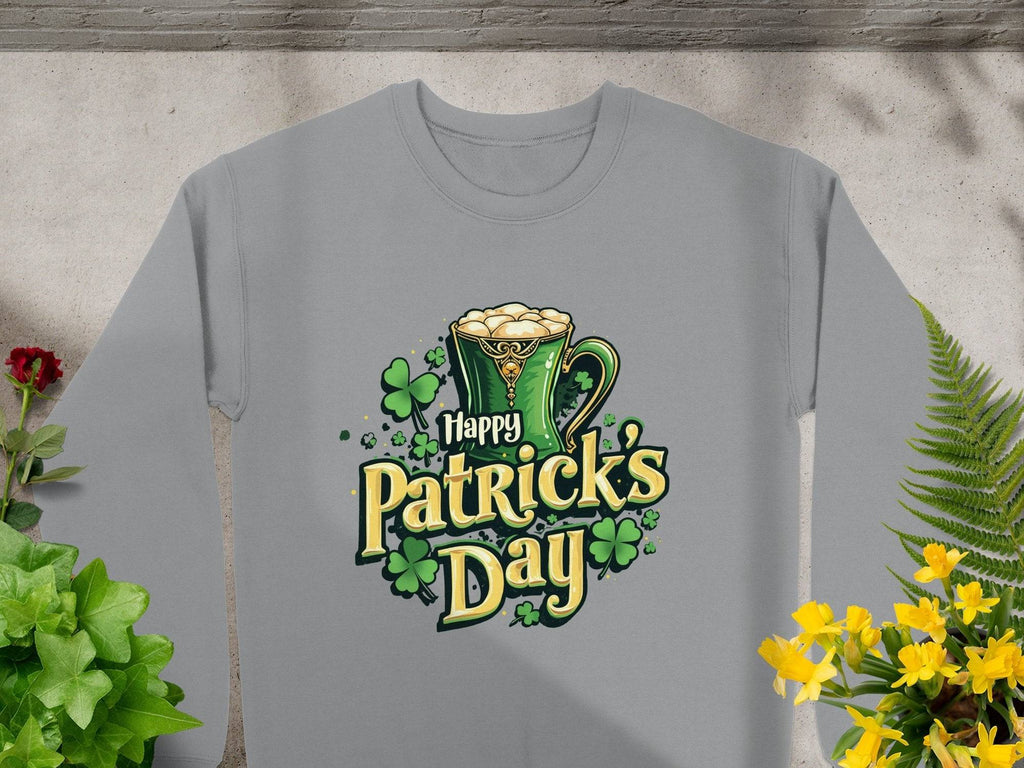The Garment Graphics sweatshirt features a festive graphic of a green mug with frothy liquid, surrounded by shamrocks and the words Happy St. Patricks Day. The gray shirt is displayed elegantly on a stone surface amidst plants.