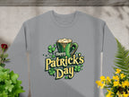 The Garment Graphics sweatshirt features a festive graphic of a green mug with frothy liquid, surrounded by shamrocks and the words Happy St. Patricks Day. The gray shirt is displayed elegantly on a stone surface amidst plants.