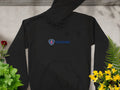 A Garment Graphics black hoodie features a blue Scania logo and red griffin emblem, displayed on concrete. Green plants and yellow flowers surround it on the right, with a single red rose adding contrast on the left.