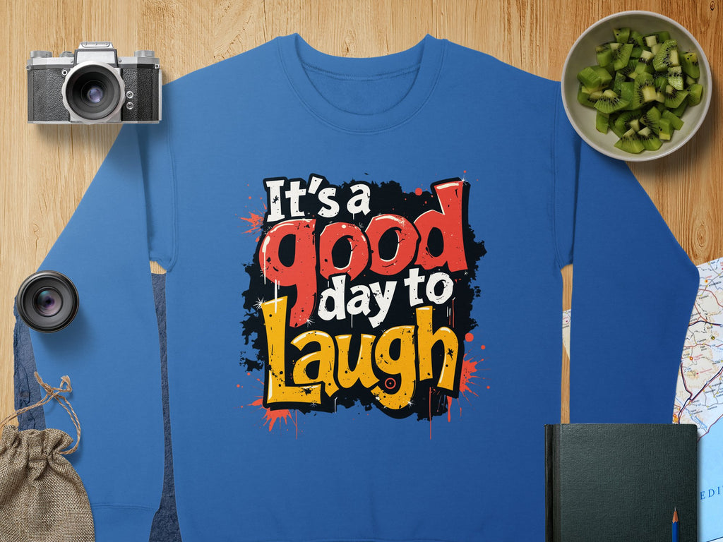 Garment Graphics offers a blue graphic sweatshirt featuring Its a good day to Laugh in bold, colorful letters. Designed for unisex sizing, its made from medium-heavy fabric. The background includes a camera, sliced cucumber jar, lens, pouch, book, and map on wood.