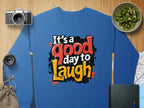 Garment Graphics offers a blue graphic sweatshirt featuring Its a good day to Laugh in bold, colorful letters. Designed for unisex sizing, its made from medium-heavy fabric. The background includes a camera, sliced cucumber jar, lens, pouch, book, and map on wood.