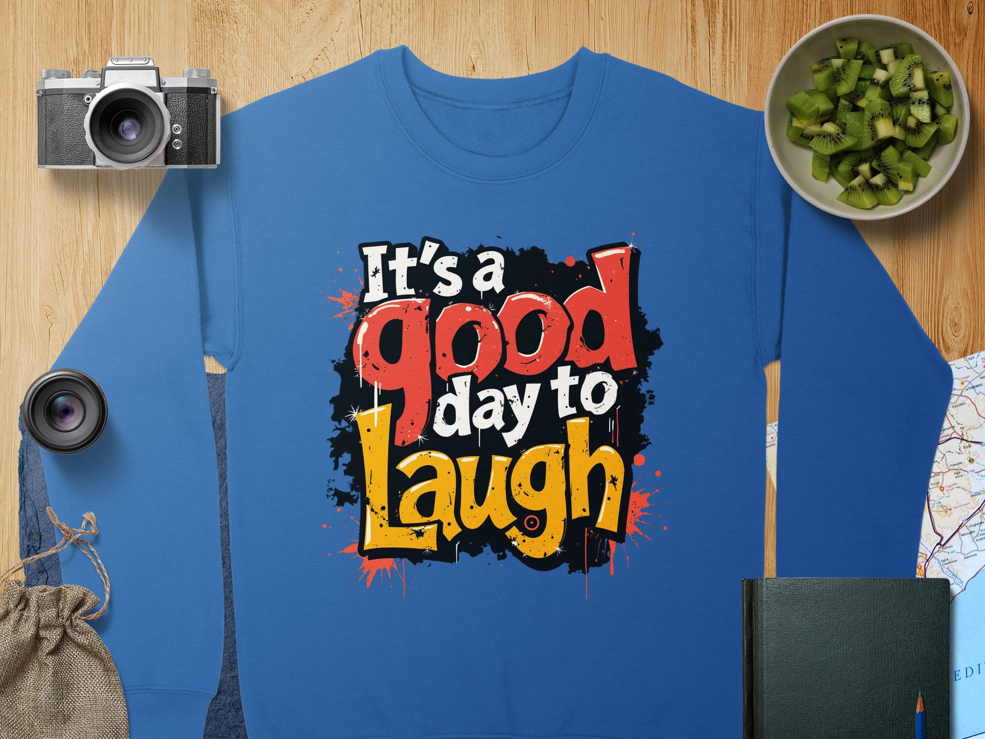 Garment Graphics offers a blue graphic sweatshirt featuring Its a good day to Laugh in bold, colorful letters. Designed for unisex sizing, its made from medium-heavy fabric. The background includes a camera, sliced cucumber jar, lens, pouch, book, and map on wood.
