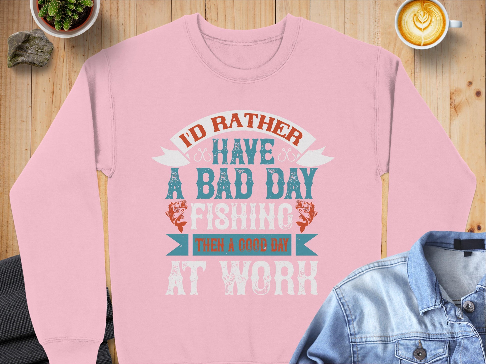 A cozy pink fishing sweatshirt from Garment Graphics reads Id rather have a bad day fishing than a good day at work. Its styled on wood with a denim jacket, rocks, a plant, and a latte. Ideal for fishing fans, its unisex sizing suits everyone.