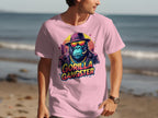 A man makes a bold statement in a Garment Graphics light pink T-shirt, featuring a vibrant Gorilla Gangster graphic with a hat and sunglasses against a sandy beach and ocean backdrop, creating an eye-catching look.