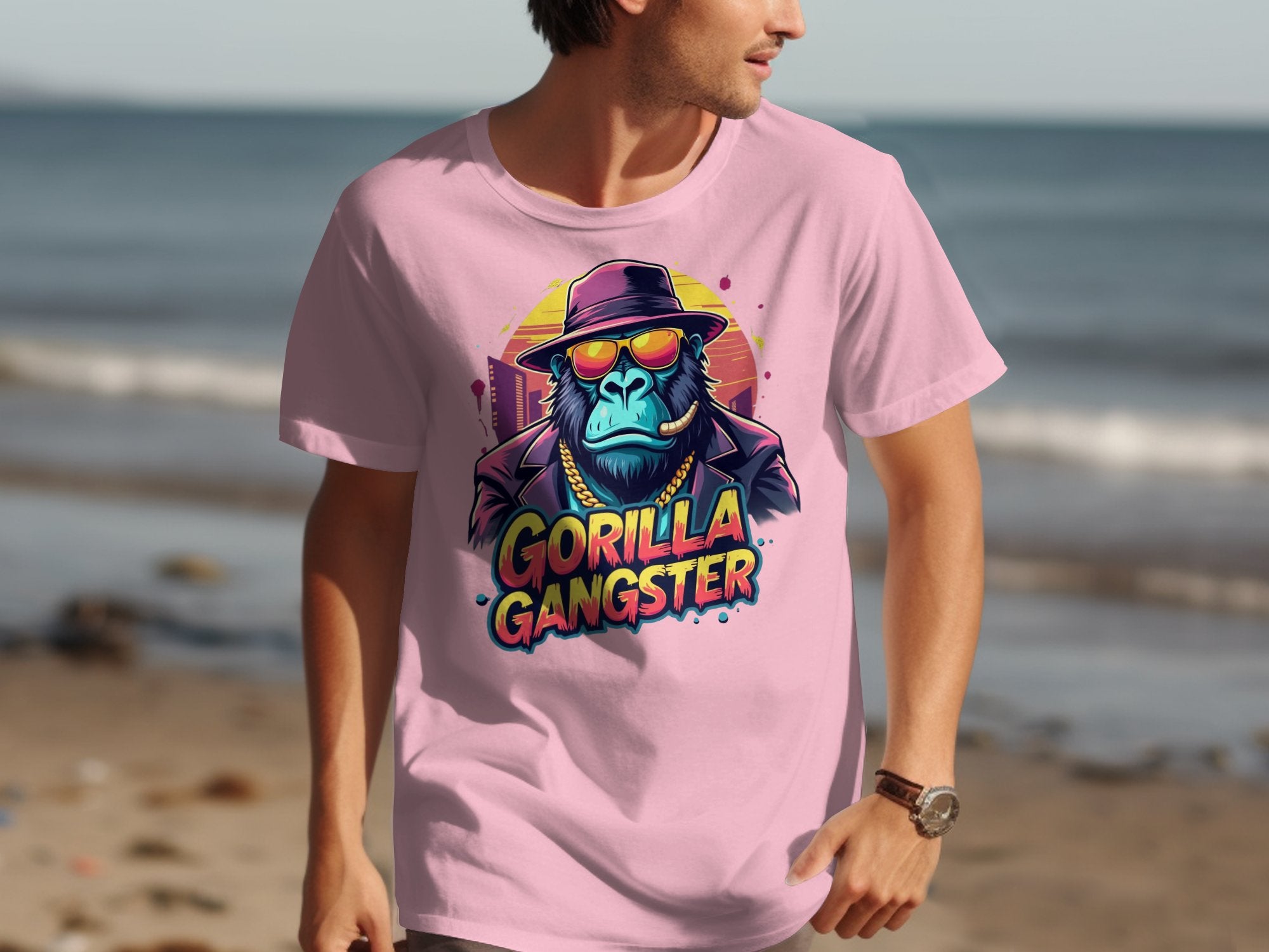 A man makes a bold statement in a Garment Graphics light pink T-shirt, featuring a vibrant Gorilla Gangster graphic with a hat and sunglasses against a sandy beach and ocean backdrop, creating an eye-catching look.