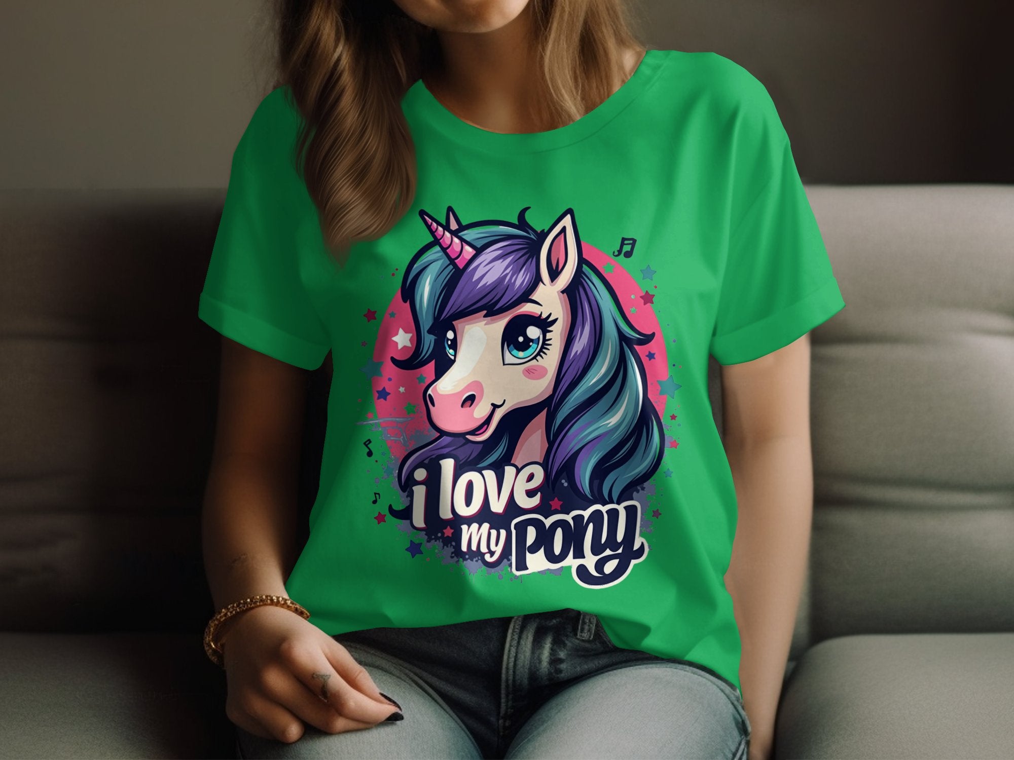 A person sits on a couch, proudly sporting a Garment Graphics I Love My Pony T-Shirt. The bright green shirt showcases an enchanting unicorn with blue and purple hair, perfect for unicorn enthusiasts. Only their lower face and hands are visible, adding an air of mystery.