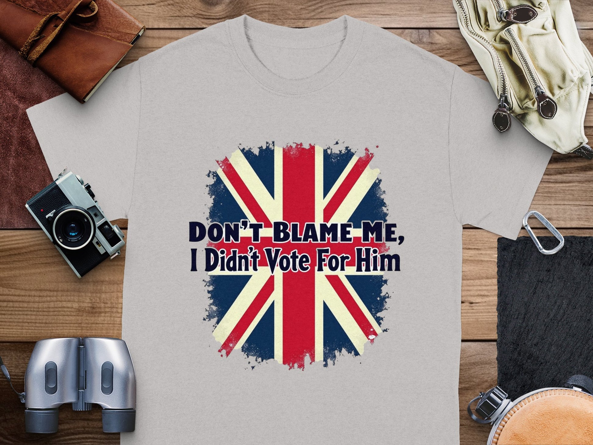 The Garment Graphics classic fit gray T-shirt features the Union Jack and the phrase Dont blame me, I didnt vote for him. Displayed on a wooden surface with binoculars, a wallet, and clothing items, it creates an intriguing visual statement.