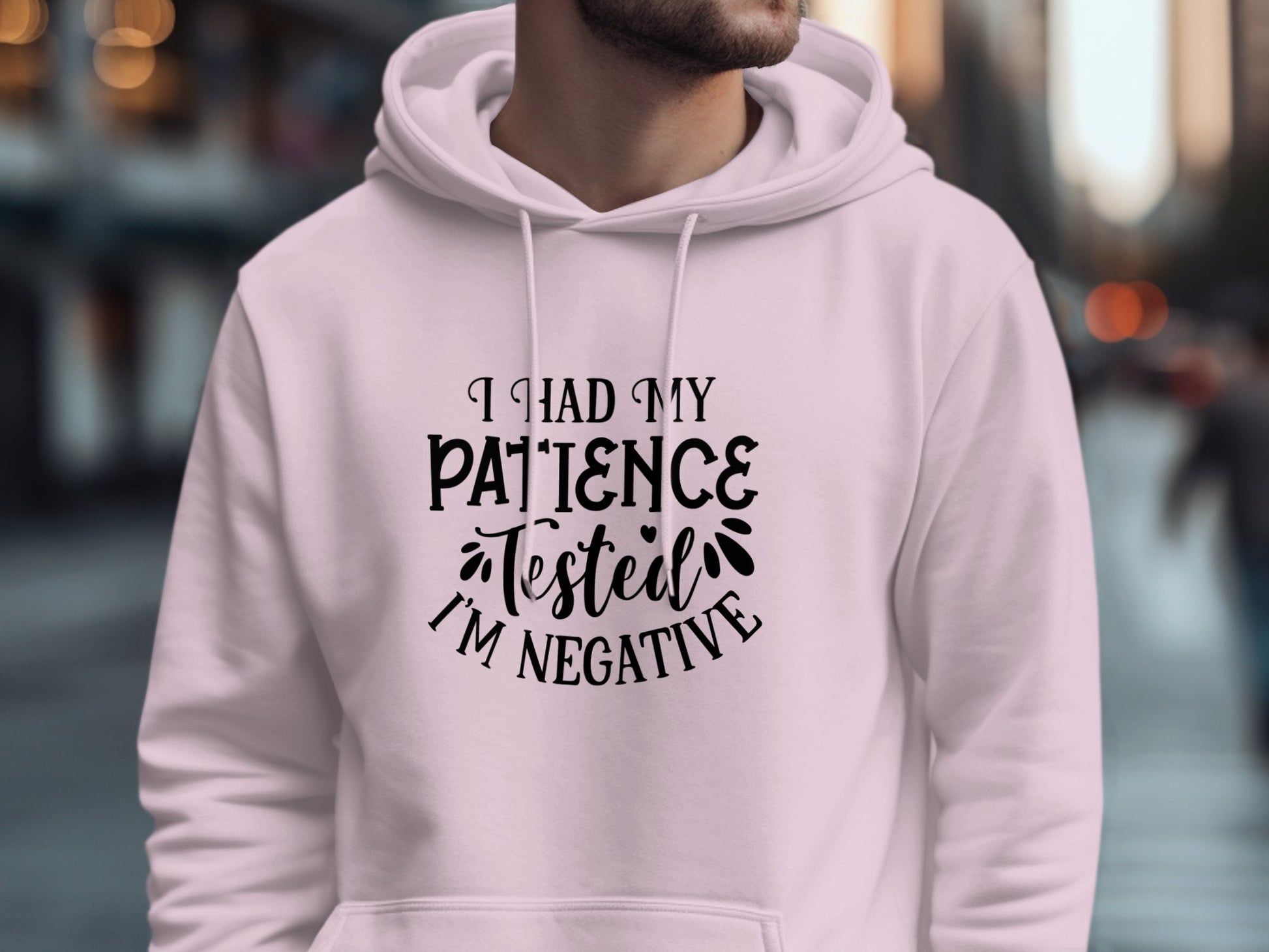 Against a blurred urban street background, a person wears a light pink hoodie by Garment Graphics, featuring the novelty graphic text I had my patience tested, Im negative in black. This unique design humorously livens up the city scene.