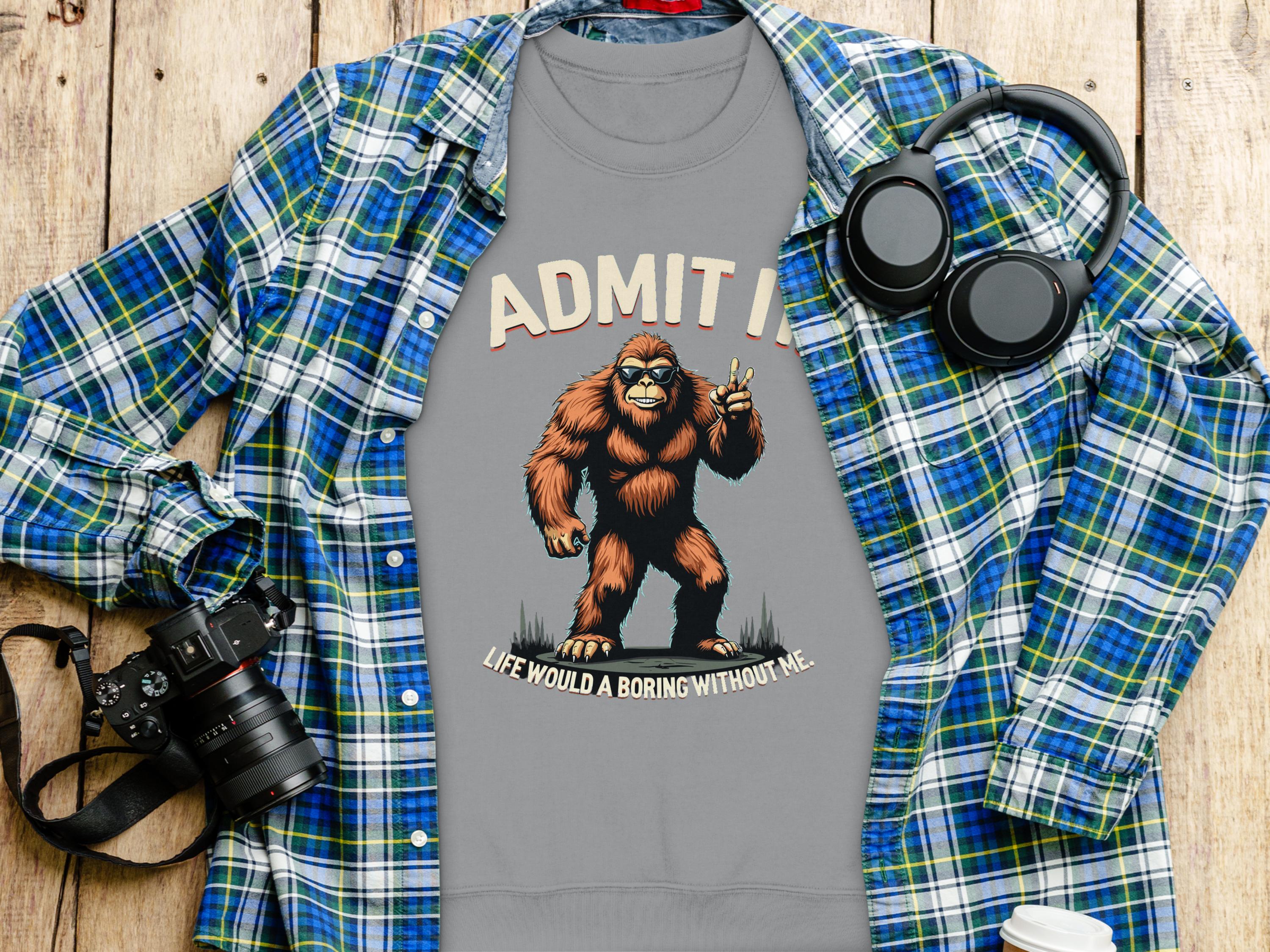 The Garment Graphics gray t-shirt features a Bigfoot graphic and the text Admit it, life would be boring without me. Its layered under a blue and green plaid shirt, with a camera, headphones, and white mug on the wooden surface nearby.